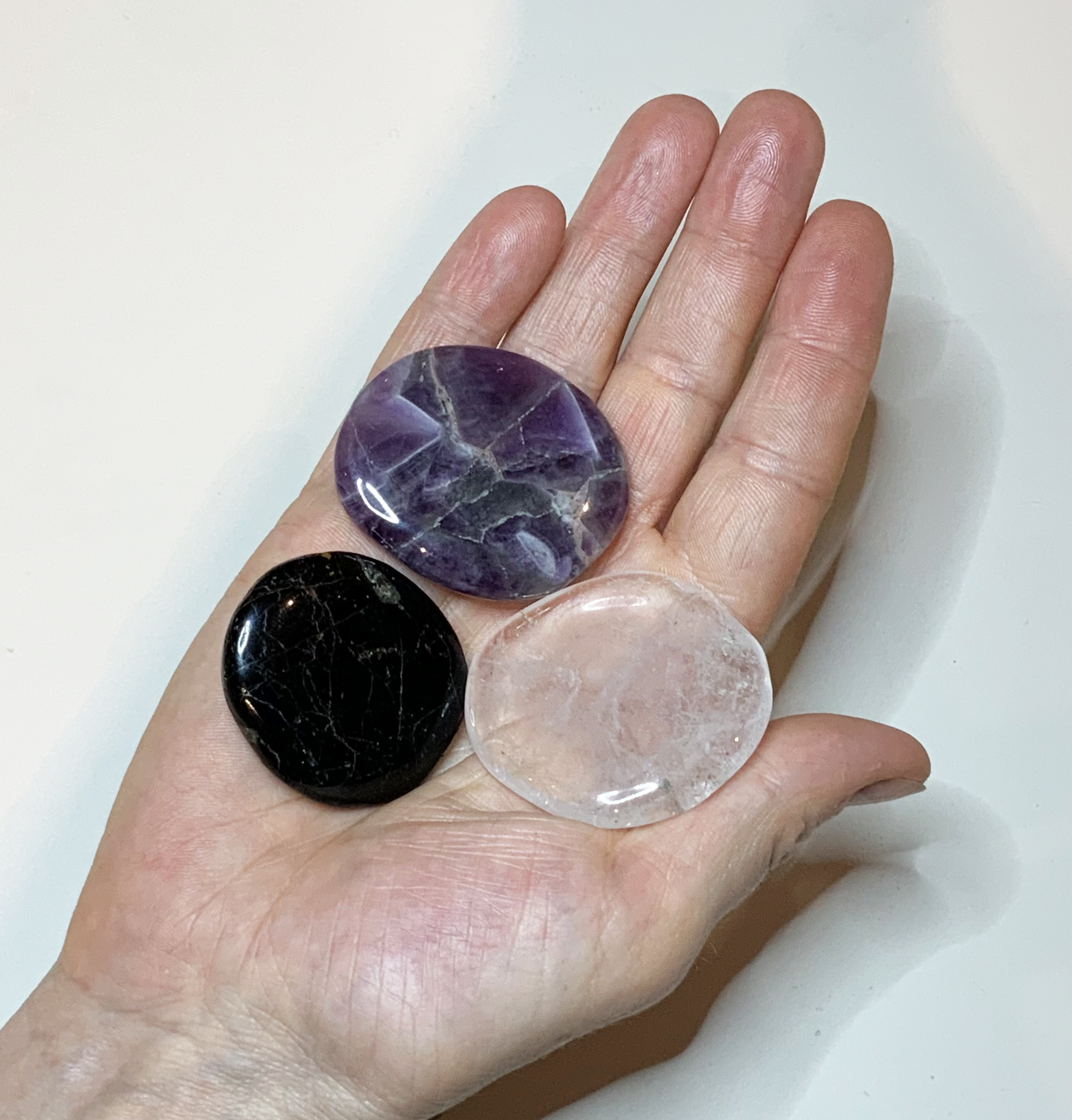 Drei polierte Schutzsteine liegen auf einer geöffneten Hand: oben ein violetter Amethyst mit hellen Maserungen, links unten ein schwarzer Schörl mit feinen Linien und rechts unten ein transparenter Bergkristall. Das Foto zeigt die Größe der Steine im Verhältnis zur Hand und ihre glatte, glänzende Oberfläche vor hellem Hintergrund.